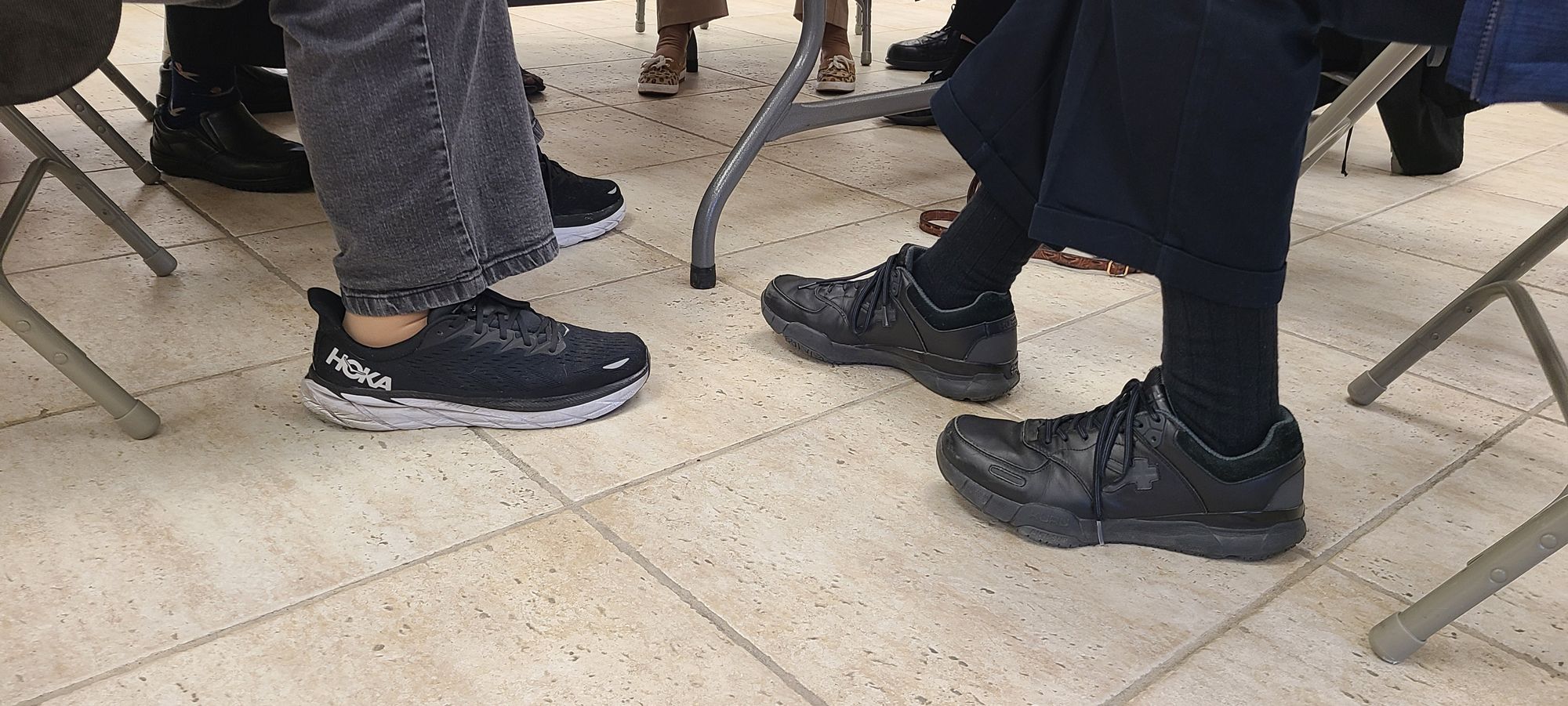 Two men sitting at the same table at the social hall of our church. One wearing black HOKAs and the other wearing black KURUs.